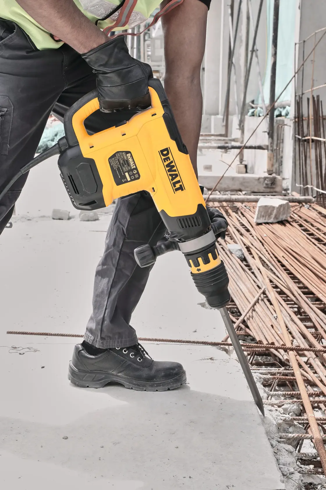 Un trabajador de construcción usando un martillo rotativo amarillo DEWALT para romper concreto en un sitio de construcción. El trabajador lleva guantes y botas protectoras.