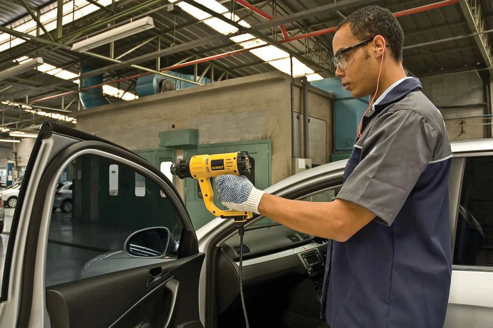 Una persona con guantes está usando una pistola de calor amarilla DEWALT (modelo D26411) en el interior de la puerta de un coche en un taller. La cara de la persona está desenfocada por privacidad.
