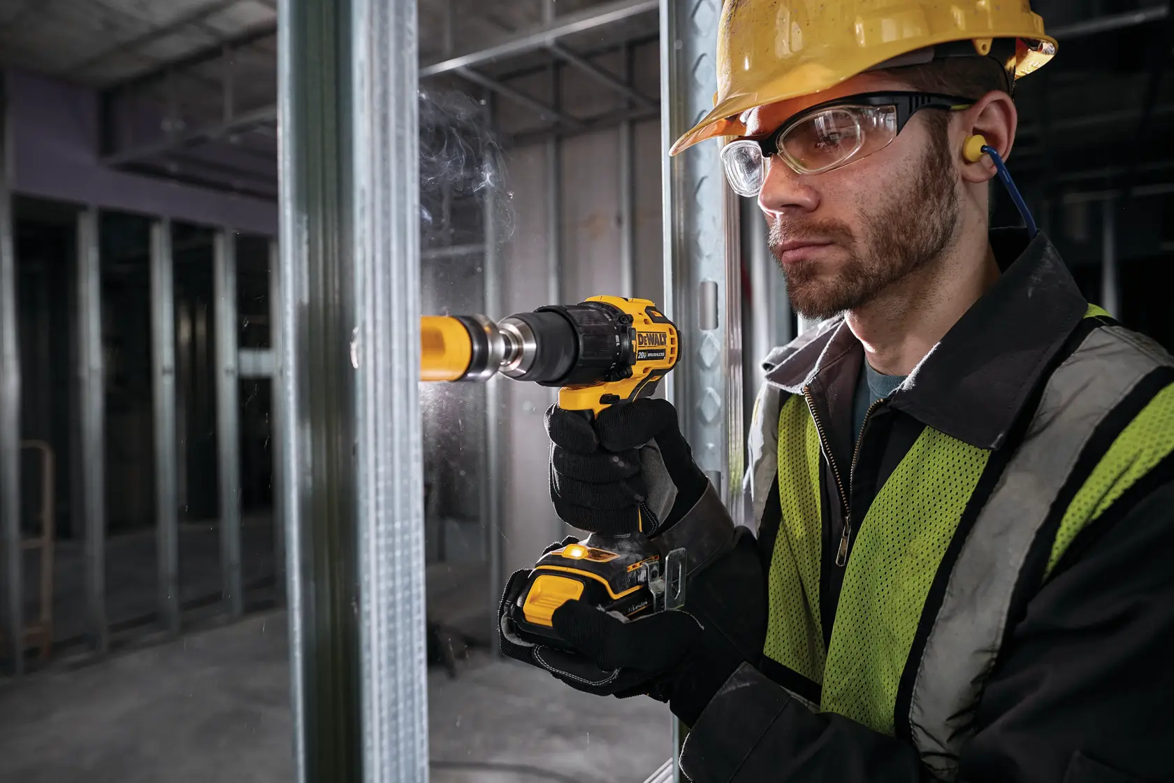 Una persona con equipo de seguridad utiliza un taladro inalámbrico DEWALT para perforar un agujero en un marco metálico en un entorno de construcción. El rostro de la persona está oculto y lleva casco, chaleco de seguridad, guantes y protección auditiva.