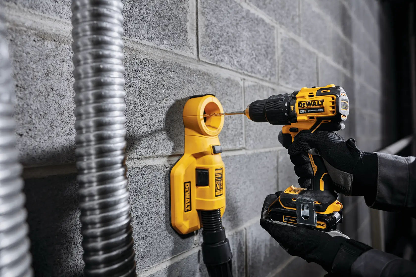 Una persona con guantes usa un taladro inalámbrico DEWALT para perforar una pared de concreto. Un accesorio amarillo de extracción de polvo DEWALT está fijado a la pared y conectado a una manguera para capturar el polvo durante la perforación.