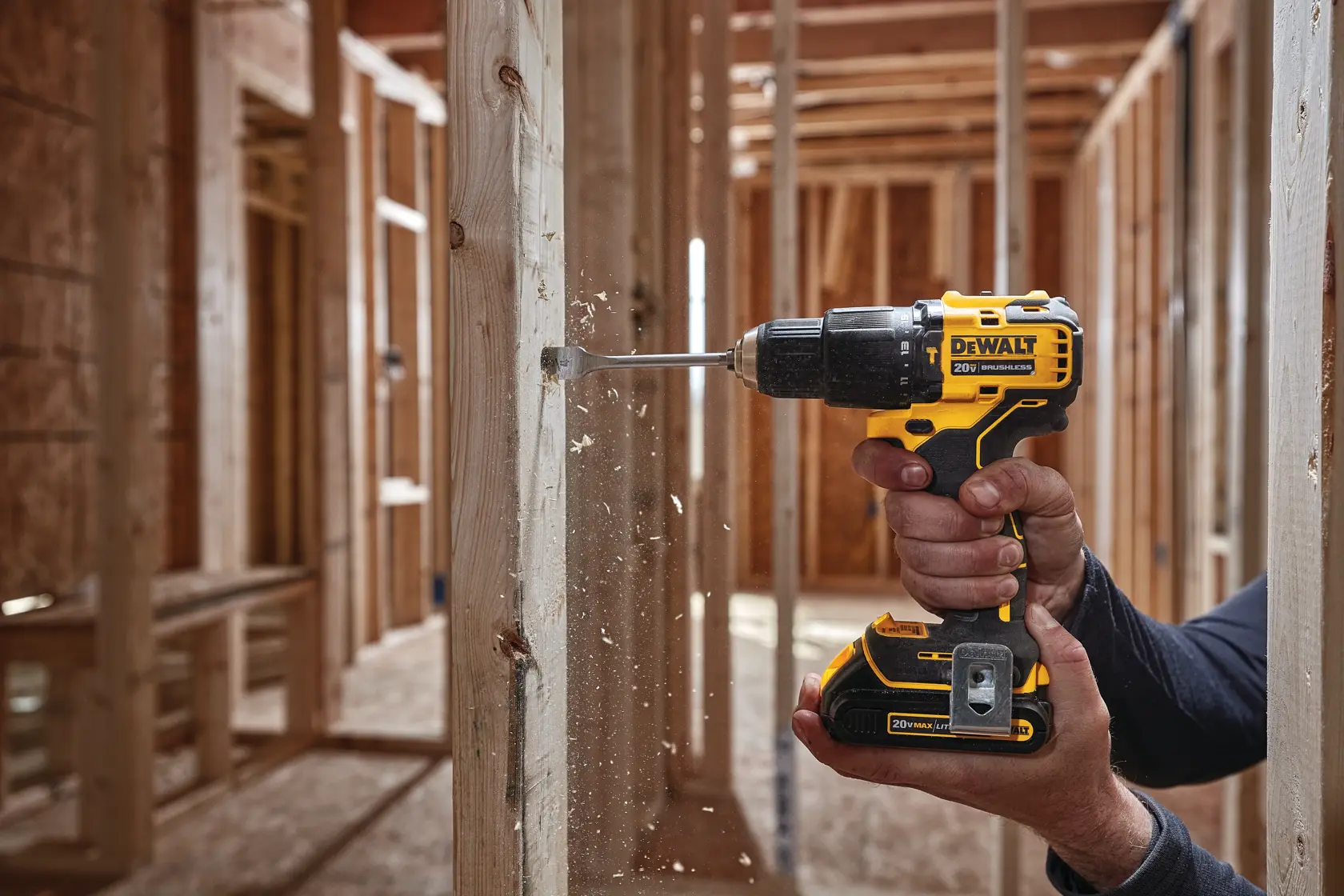 Una persona utiliza un taladro inalámbrico amarillo y negro DEWALT para perforar un agujero en una viga de madera en una obra de construcción, con virutas de madera saliendo de la broca.
