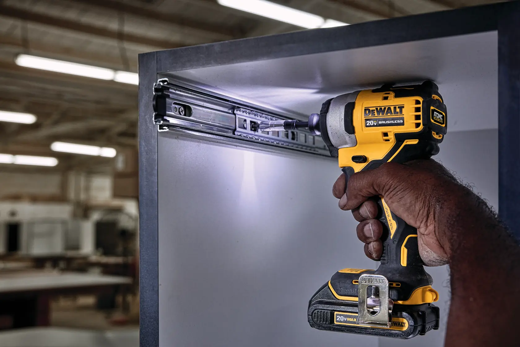 Una persona usa un atornillador de impacto inalámbrico DEWALT de 20V para instalar o ajustar una guía metálica para cajón dentro de un mueble, en un taller.