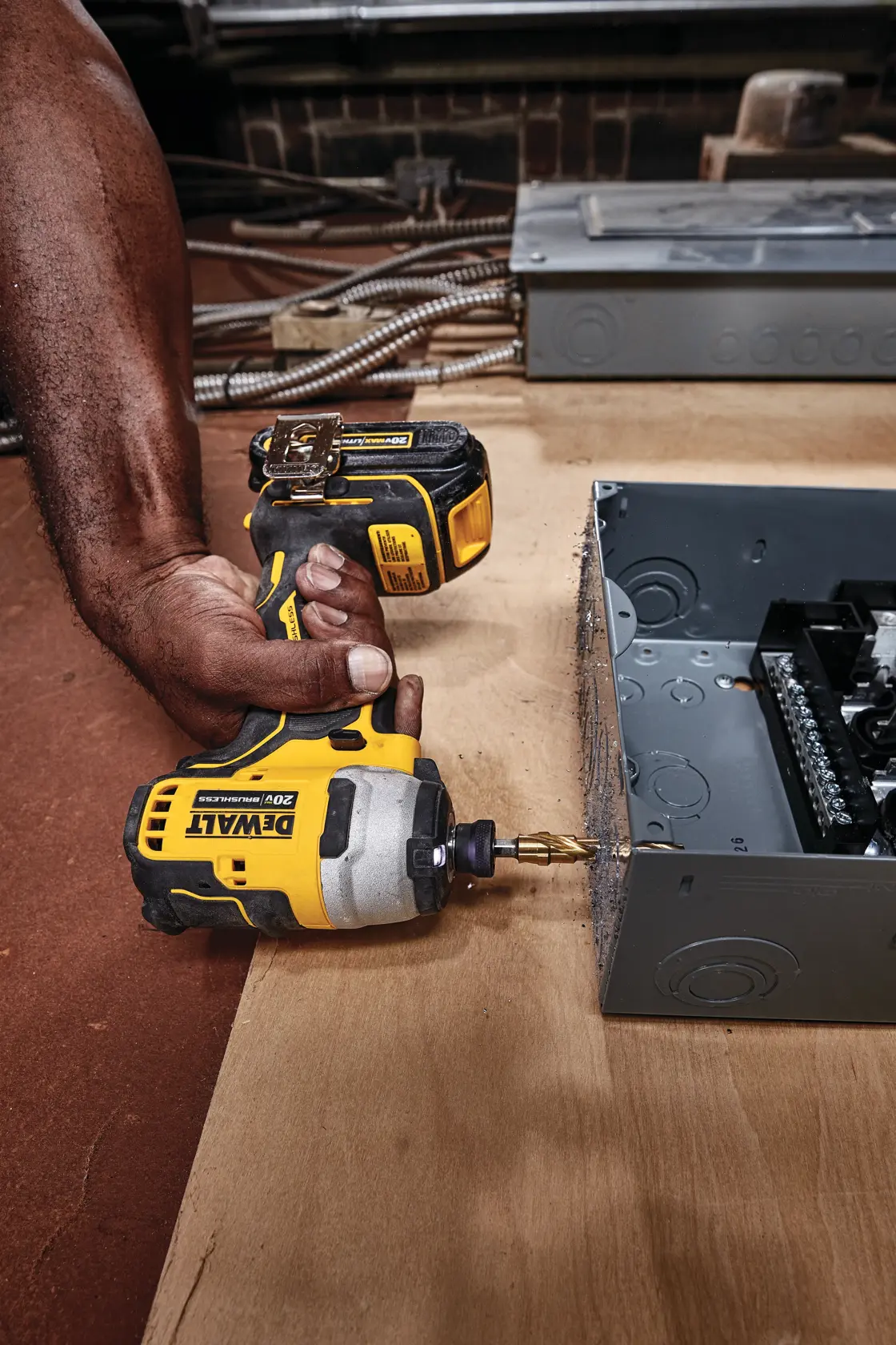 Una persona está usando un destornillador de impacto inalámbrico DEWALT DCF809B de 20V para perforar una caja eléctrica de metal sobre una superficie de madera.