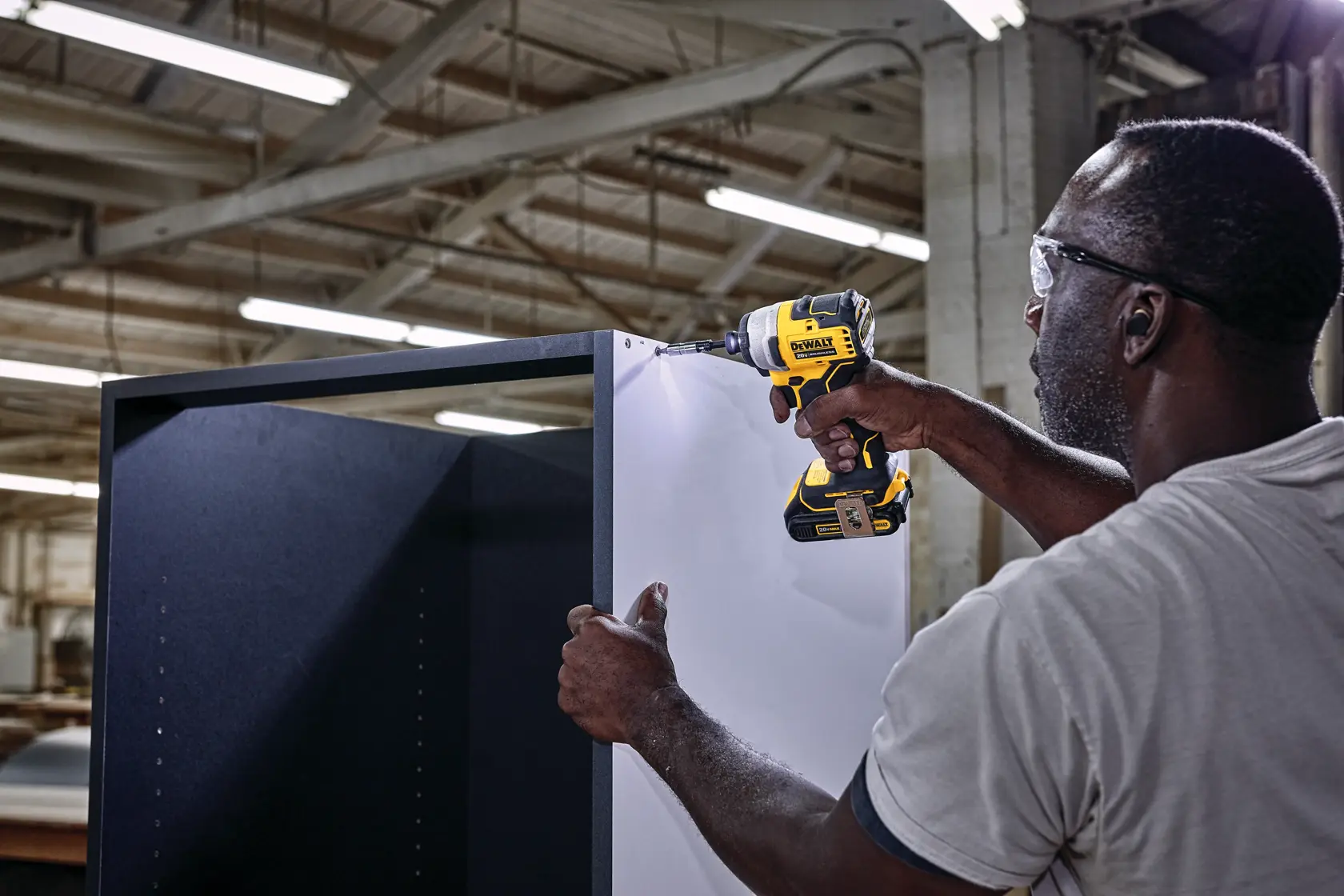 Una persona utiliza un taladro inalámbrico DEWALT, modelo DCF809B, para ensamblar un gabinete grande dentro de un taller industrial con luces y vigas visibles arriba.