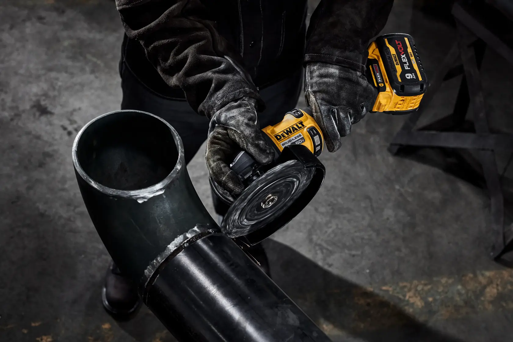Una persona con guantes protectores utiliza una amoladora inalámbrica DEWALT para cortar o pulir una tubería metálica negra grande en un entorno industrial.