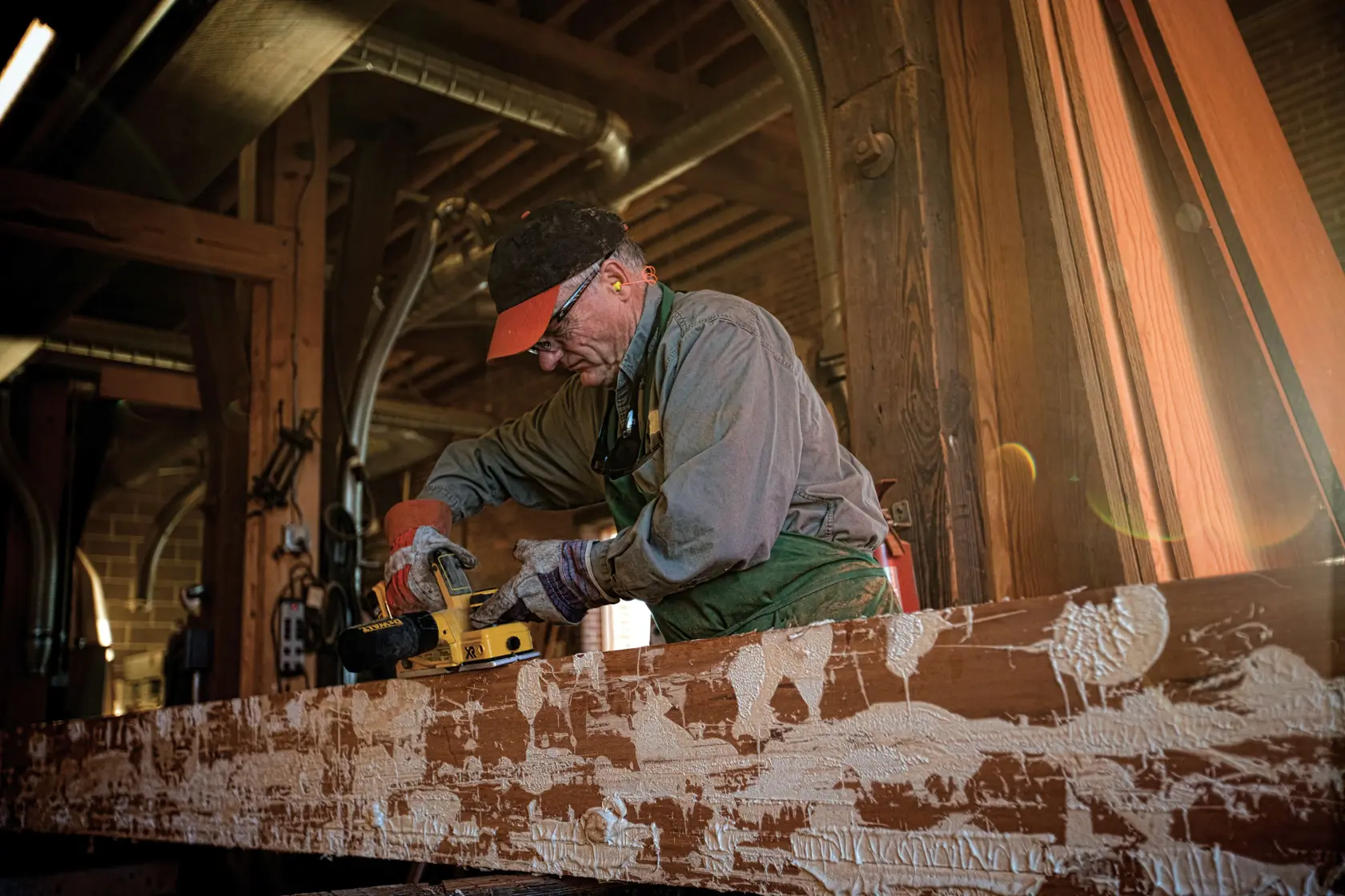Una persona usando una cepilladora inalámbrica DEWALT DCP580B sobre una viga de madera en un taller. El individuo lleva guantes y ropa de trabajo. Al fondo se ven equipos de taller y materiales de madera.