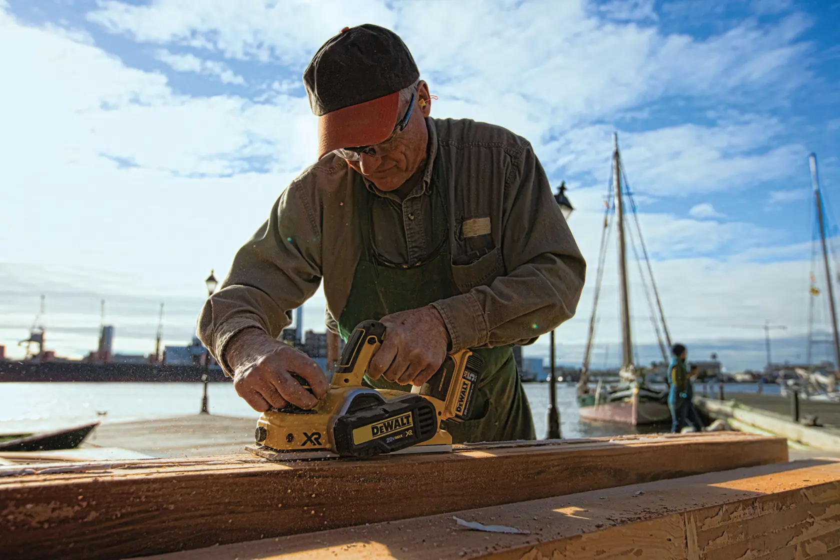 Una persona está utilizando una cepilladora inalámbrica DEWALT DCP580B para alisar una pieza de madera al aire libre cerca de un puerto deportivo, con barcos y agua visibles en el fondo. El rostro de la persona está oculto.