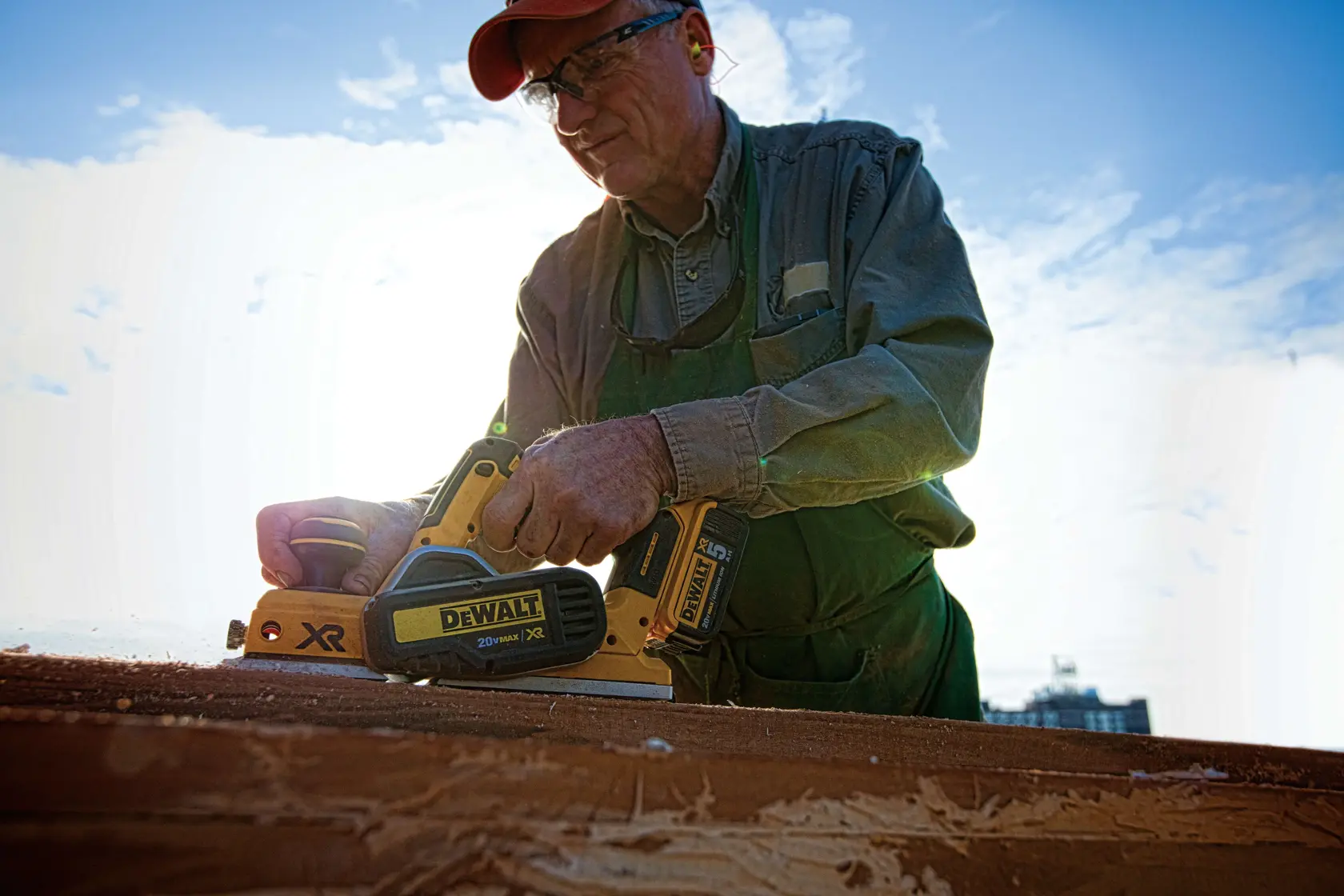 Una persona usando una cepilladora inalámbrica DEWALT DCP580B, trabajando en un trozo de madera al aire libre bajo un cielo parcialmente nublado. El rostro de la persona está difuminado por privacidad.