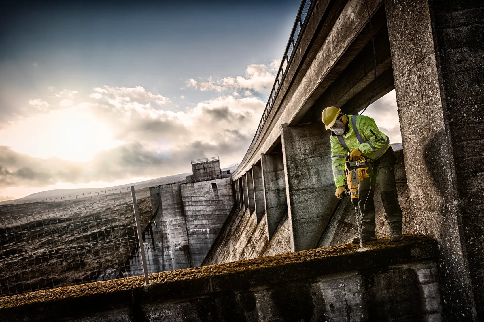 Trabajador usa taladro DEWALT en el concreto junto a una gran estructura industrial al aire libre.