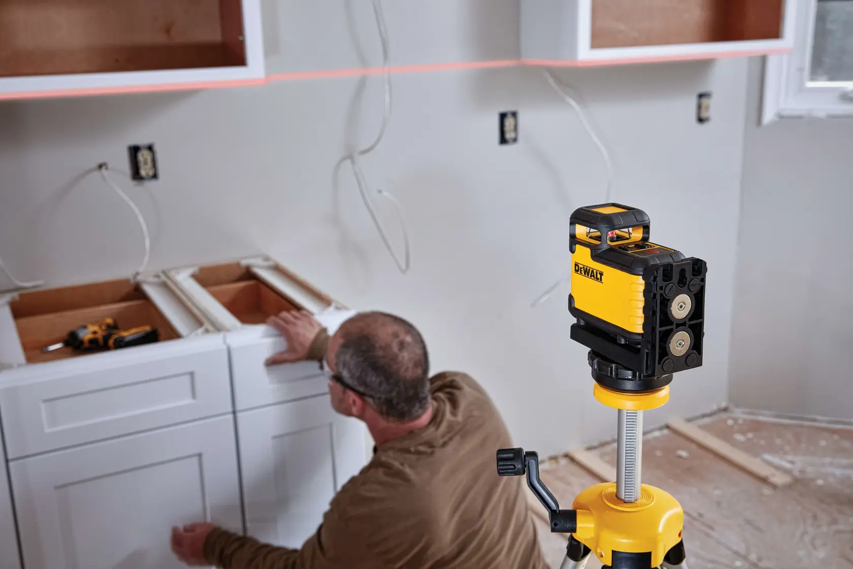 Un nivel láser DEWALT está instalado en un trípode en una cocina en construcción. Una persona está instalando gabinetes blancos y el láser proyecta una línea recta en la pared para alineación.