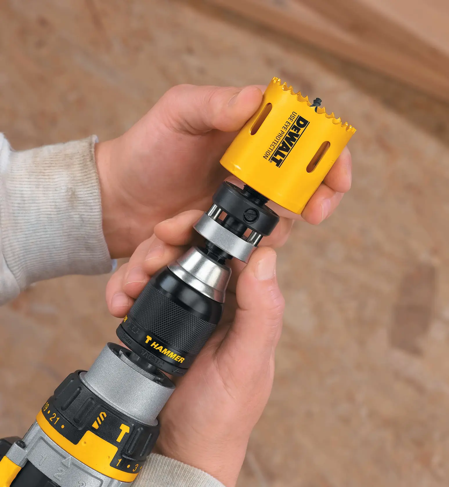 Primer plano de manos colocando una sierra de corona amarilla DEWALT en un taladro con un adaptador de cambio rápido, con un fondo desenfocado.