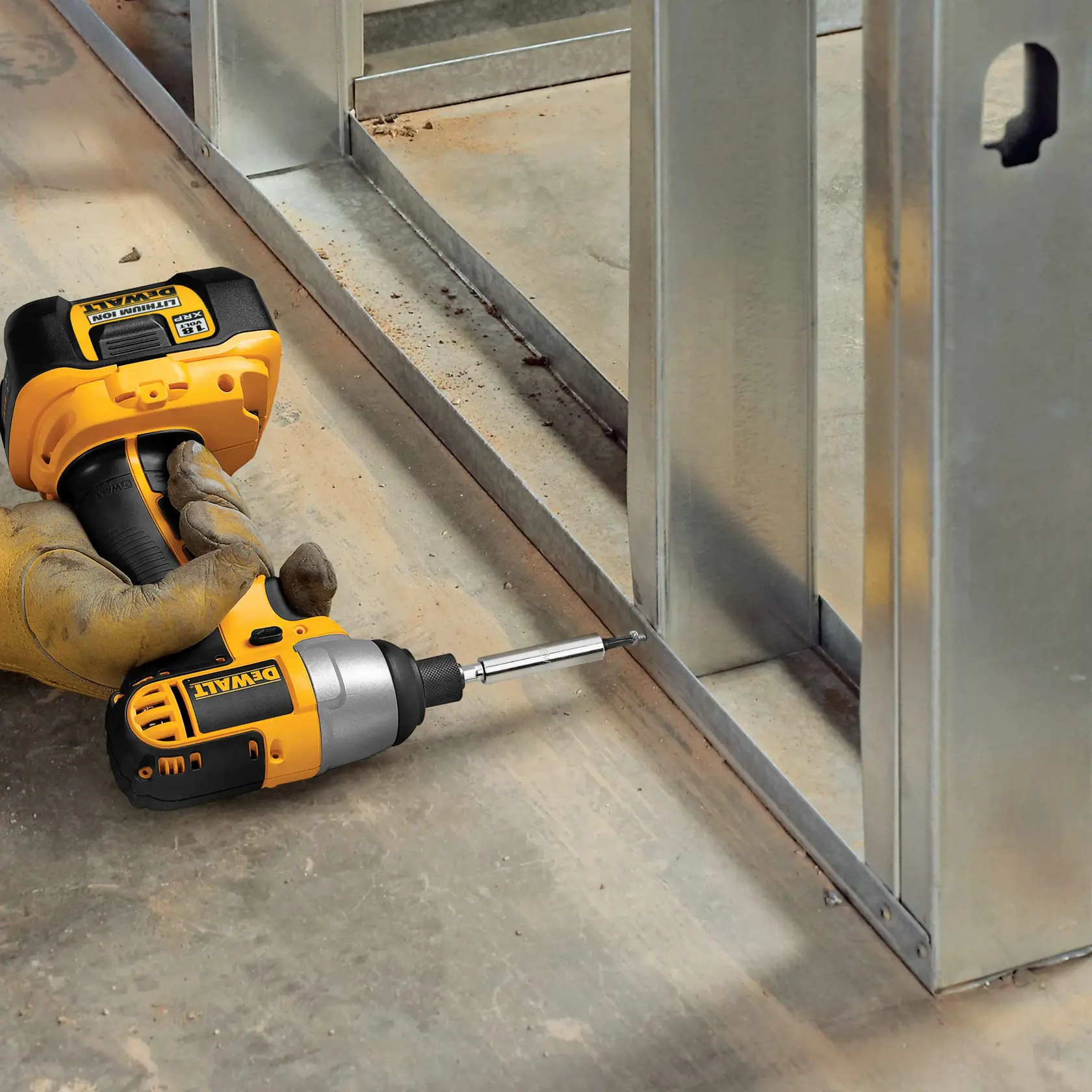 Una persona con un guante de trabajo utiliza un taladro inalámbrico DEWALT con una punta de destornillador para fijar un tornillo en una estructura de metal sobre un suelo de concreto.