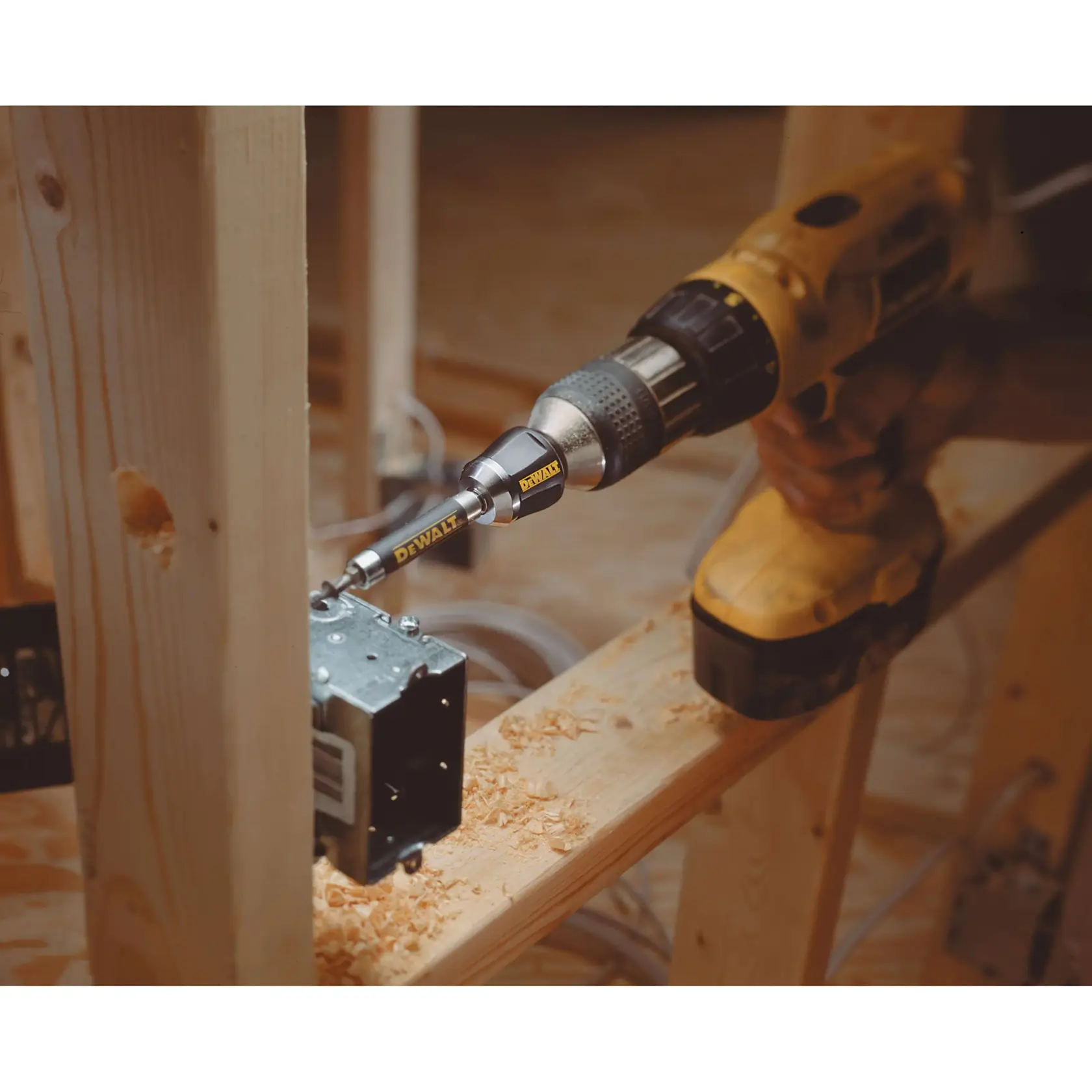 Un taladro DEWALT con una punta DEWALT está siendo utilizado para atornillar tornillos en una caja eléctrica de metal montada en un marco de madera. Hay virutas de madera sobre la viga y el entorno parece ser un sitio de construcción interior.