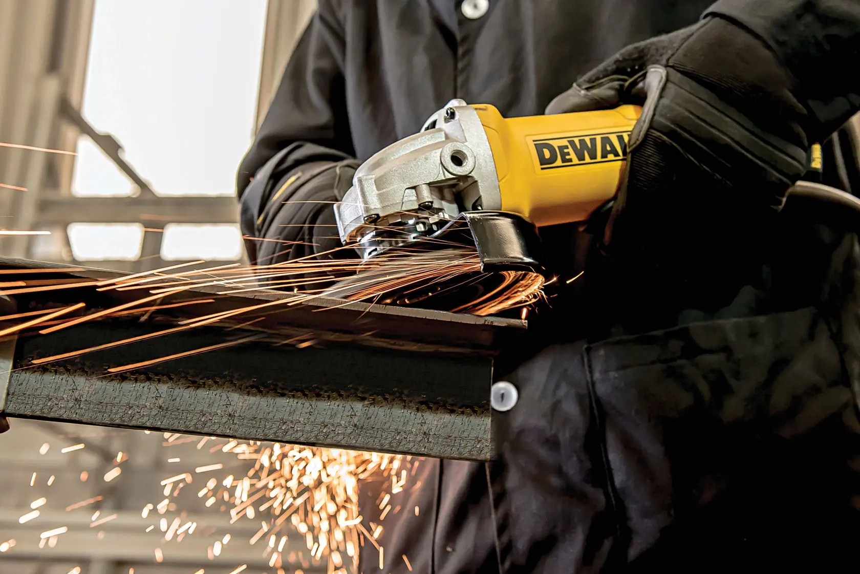 Primer plano de una persona con guantes protectores y chaqueta usando una amoladora angular amarilla DEWALT para cortar una pieza de metal, con chispas volando.