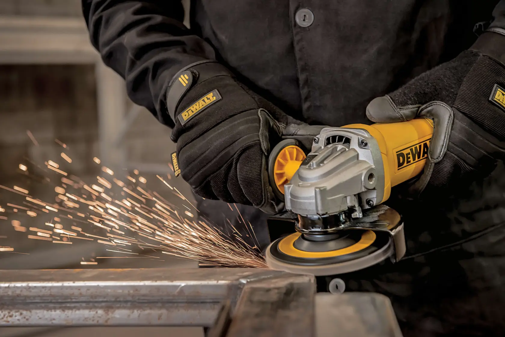 Una persona con guantes protectores negros y chaqueta está usando una amoladora angular amarilla DEWALT para cortar una pieza de metal, lo que provoca chispas.