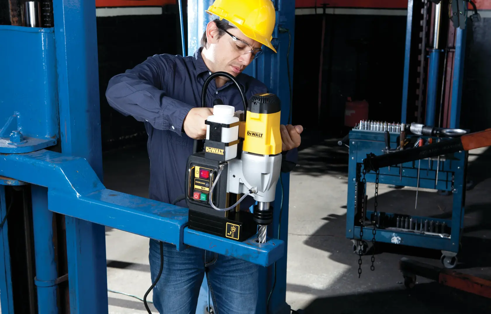 Una persona con casco amarillo opera una taladradora magnética DEWALT DWE1622K sobre una viga de metal azul en un entorno de taller industrial. El rostro de la persona está difuminado por privacidad.