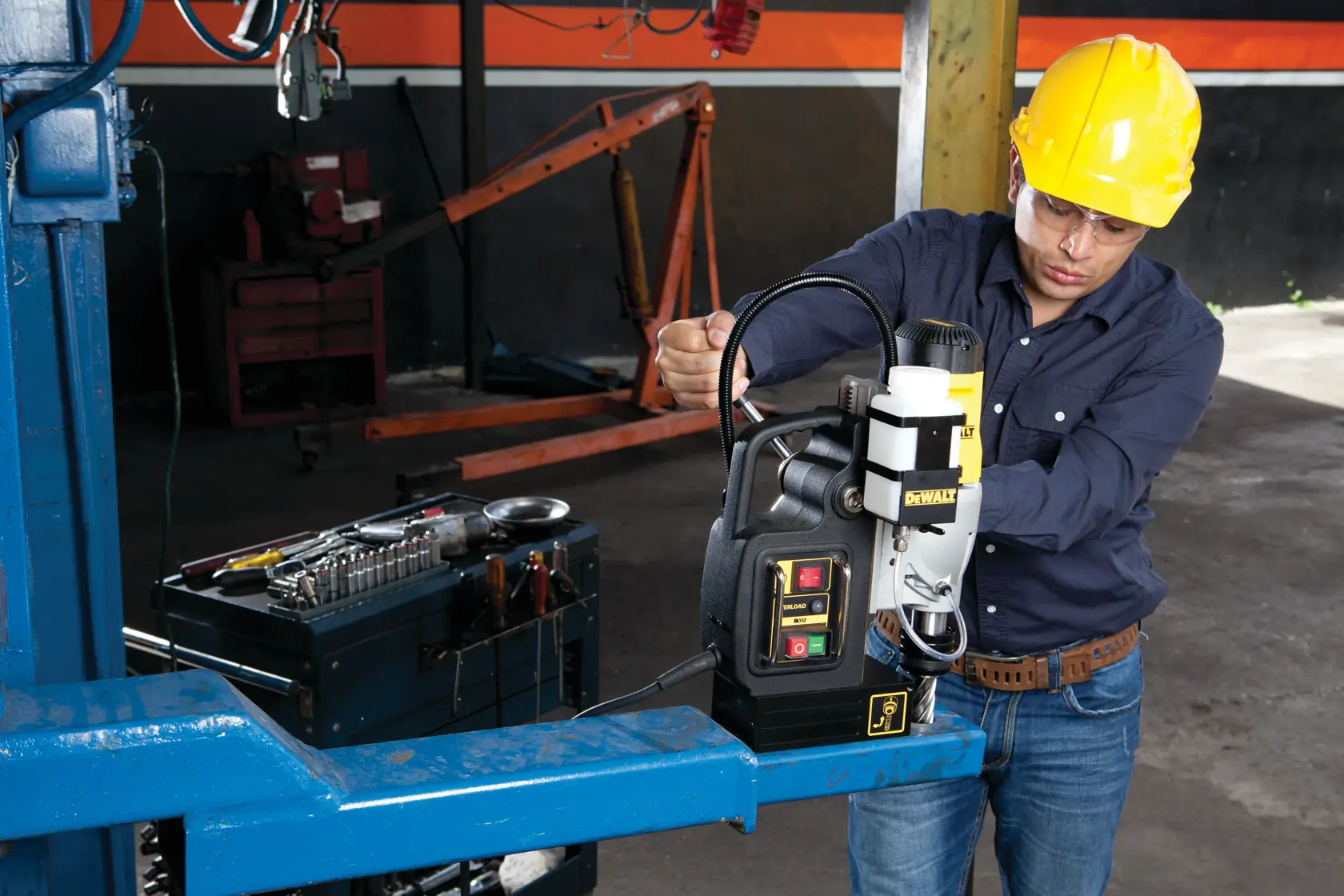 Una persona con un casco de seguridad amarillo opera un taladro magnético DEWALT DWE1622K en una viga de metal en un taller industrial. Cerca hay una caja de herramientas con varios utensilios. El rostro de la persona está difuminado para proteger su privacidad.