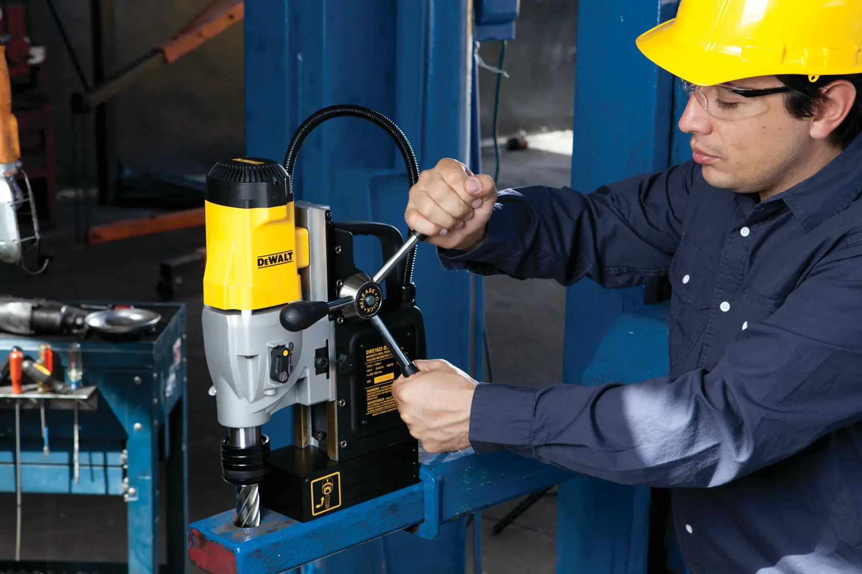 Una persona con casco amarillo opera una prensa taladradora magnética DEWALT, modelo DWE1622K, montada sobre una viga de metal azul en un taller industrial. El rostro de la persona está difuminado por privacidad.